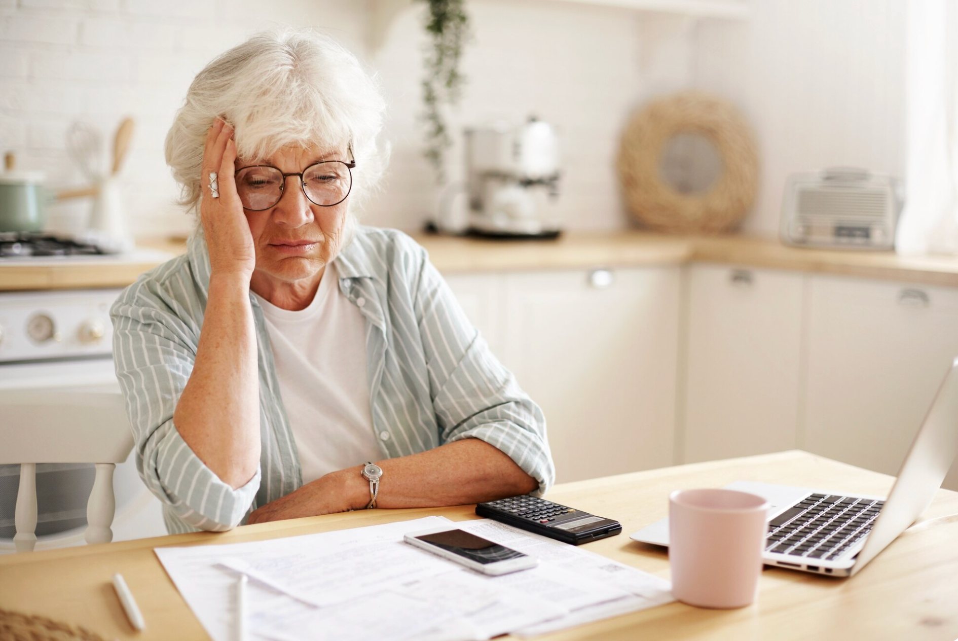 An older woman at her kitchen table, head in hand, reviewing bills with a calculator and laptop