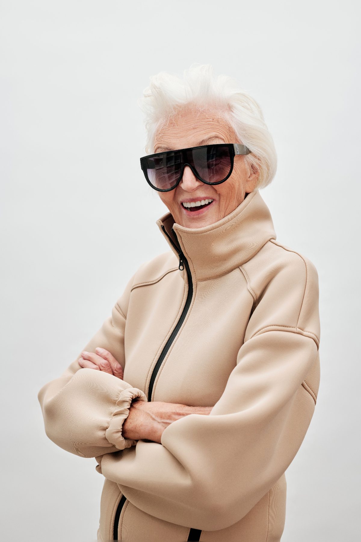 Woman with white hair in oversized sunglasses and a camel zip jacket, laughing